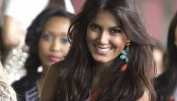 A woman with long dark hair and colorful earrings smiles at the camera. She is in focus, while another woman in the background appears blurred.