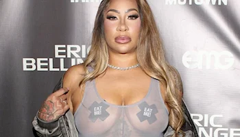 A woman with long, wavy hair poses in a sheer top with two X-shaped stickers that read EAT ME covering her chest. She stands in front of a backdrop with text.