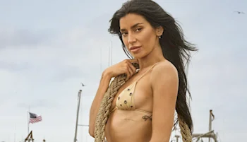 A woman with long dark hair wearing a studded bikini holds a thick rope over her shoulder, standing outdoors with a cloudy sky and an American flag in the background.