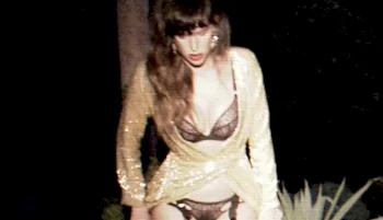A woman in lingerie and a gold robe poses outdoors at night next to plants.