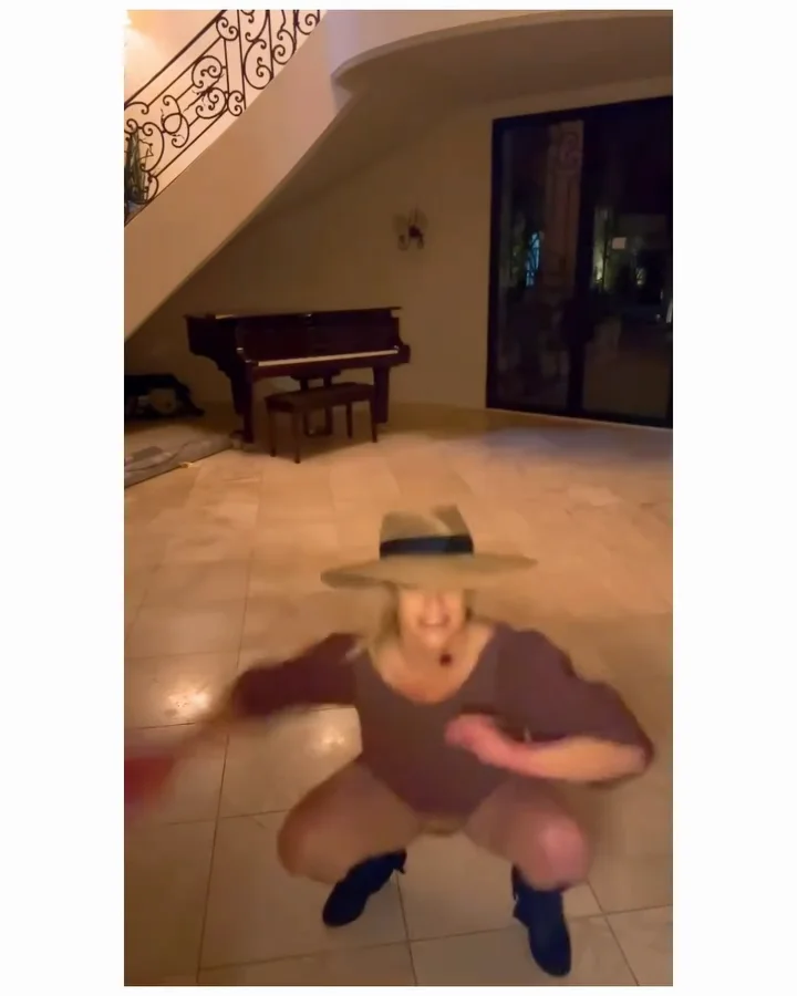 Britney Spears wearing a hat, long-sleeve top, and boots squats energetically indoors on a tiled floor, with a piano and a staircase visible in the background.