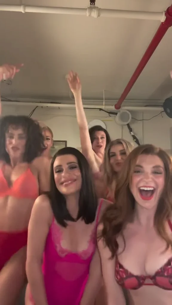 Six women in colorful lingerie smile and pose energetically in a room with exposed pipes and a speaker on the wall, some raising their arms and dancing.