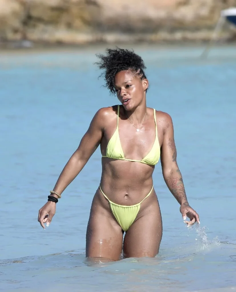 A woman wearing a light green bikini stands in shallow, clear blue water at the beach, with rocks visible in the background. She has short curly hair and several tattoos on her arms.