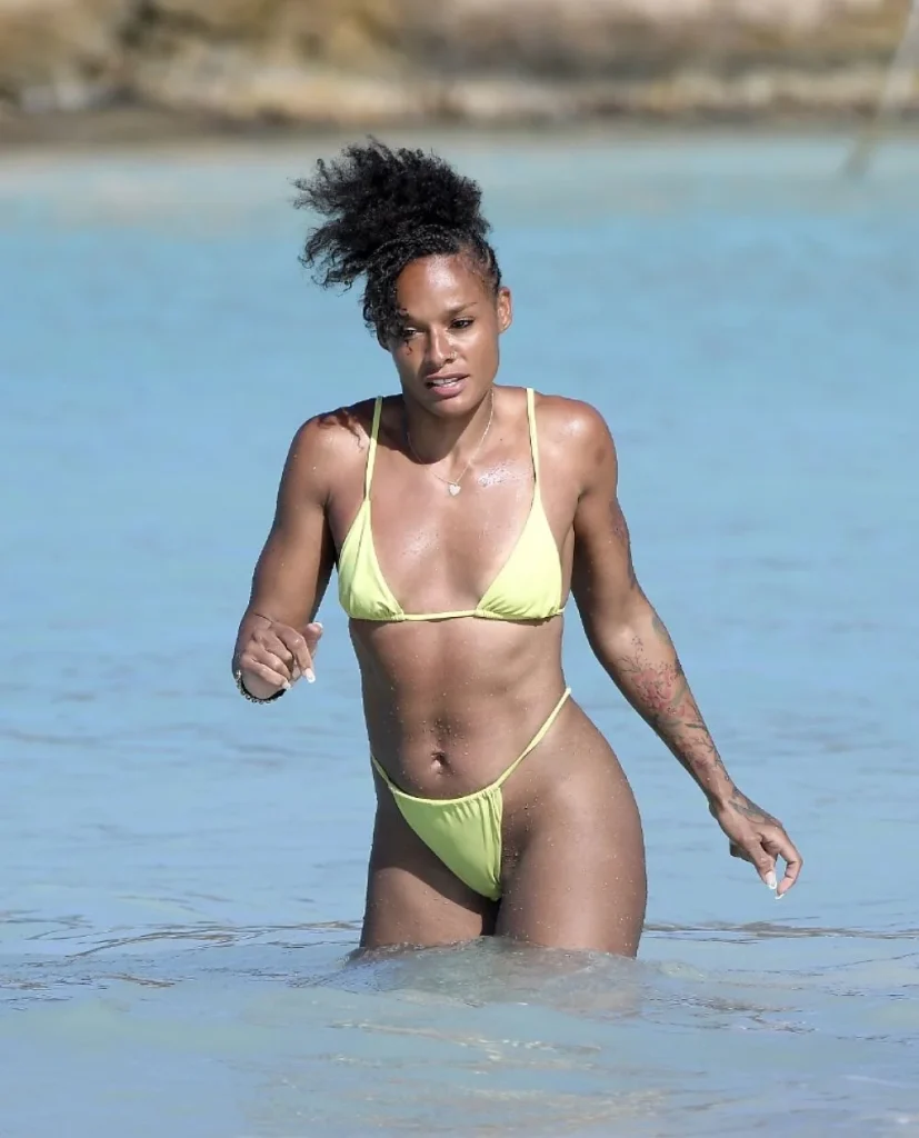 A woman wearing a yellow bikini stands in clear, shallow water at the beach. She has a tattoo on her left forearm and her hair is styled in a high, curly ponytail. The background shows more water and a blurred shoreline.