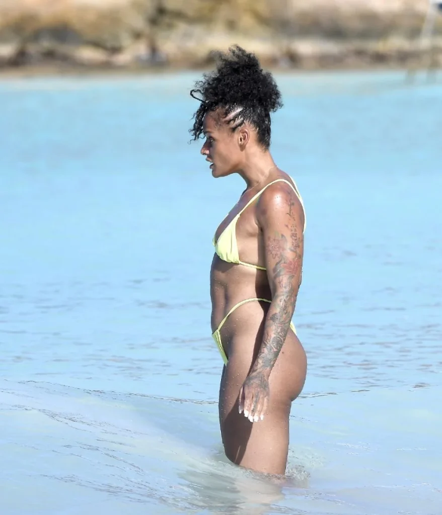 A woman with curly hair and tattoos on her arm stands in shallow ocean water, wearing a yellow bikini. The background shows blurred sandy rocks and blue water.