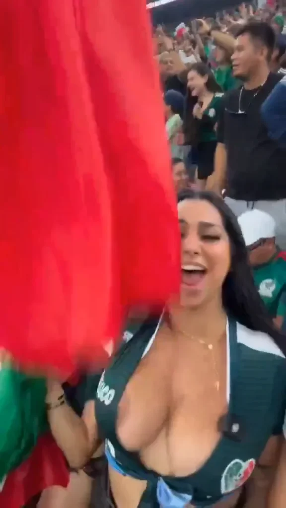 A woman wearing a green sports jersey cheers and waves a red flag in a crowded stadium, surrounded by other fans dressed in green.