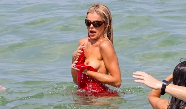 A woman in sunglasses is standing in the ocean, holding up her red swimsuit top with a surprised expression because her breast is exposed.