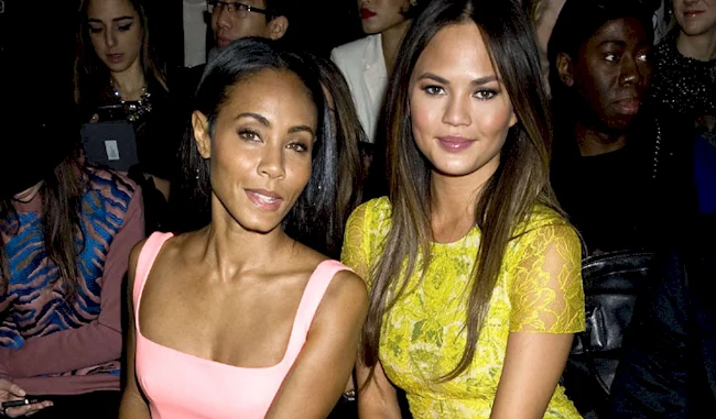 Two women sit side by side at a fashion show, dressed elegantly; one wears a sleeveless pink dress, the other a yellow lace dress. People sit behind them, some looking toward the camera.