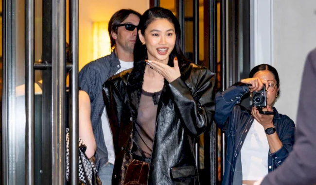 Asian woman wearing a black leather jacket and sheer top smiles and gestures while walking out of a building, followed by a man in sunglasses and another person taking a photo with a camera.