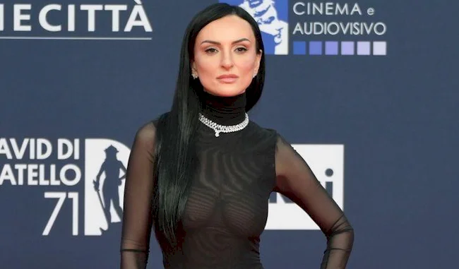 A woman with long black hair wearing a sheer black dress and a silver necklace poses confidently on a red carpet in front of a backdrop with logos and text.