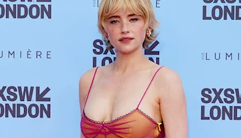A woman with short blond hair poses in front of a blue SXSW London event backdrop, wearing a sheer pink dress with thin straps and decorative accents.