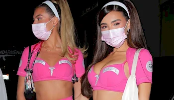 Two women wearing matching pink crop tops, skirts, and white headbands, styled as nurse costumes with medical symbols and badges, both wearing light pink face masks, standing next to each other at night.