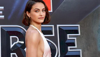 A woman with short brown hair, wearing a sleeveless, light-colored dress, poses sideways on a red carpet in front of a large sign with bold letters.