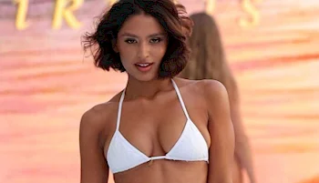 A woman with short dark hair wearing a white bikini top stands confidently in front of a blurred sunset or beach backdrop.