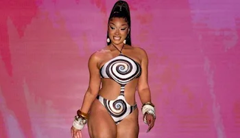 A woman walks confidently on a runway wearing a black and white spiral-patterned cutout bodysuit, accessorized with bangles and earrings, against a pink background.