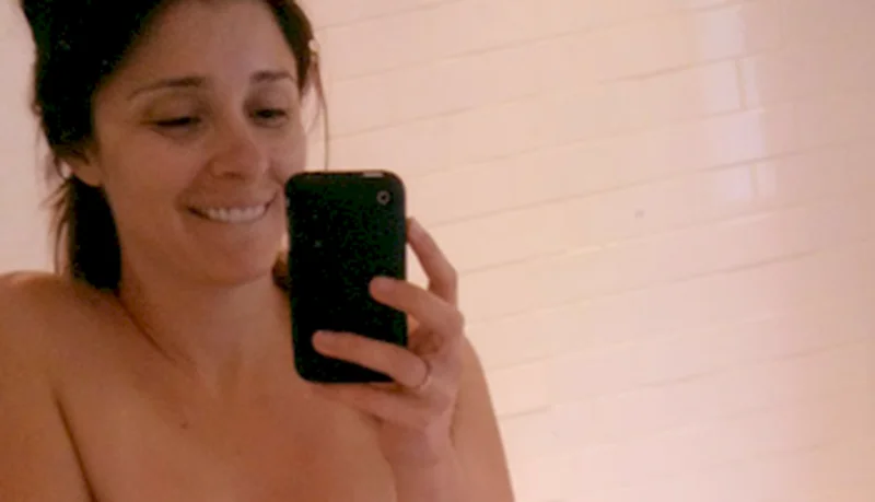 A woman takes a selfie in front of a white tiled wall, smiling and holding a smartphone.