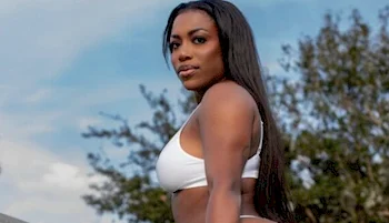 A black woman with long dark hair poses outdoors in a white bikini. There are trees and a blue sky with some clouds in the background.