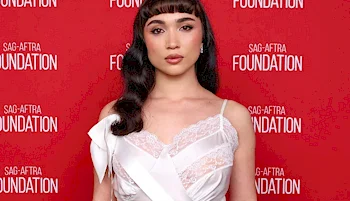 A woman wearing a white lace dress poses in front of a red backdrop with repeated white SAG-AFTRA Foundation logos.