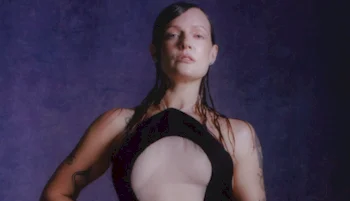 A woman with wet, slicked-back hair poses confidently against a dark purple background, wearing a black outfit with a large cutout in the chest area.