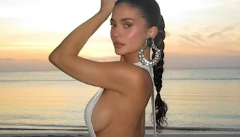 A woman with long dark hair in a braid poses on a beach at sunset, wearing a white swimsuit and large hoop earrings, with the calm ocean and colorful sky in the background.
