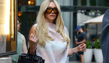 A woman with long blonde hair and large dark sunglasses is walking outside in an urban area, holding a black handbag and wearing a light-colored sleeveless top.