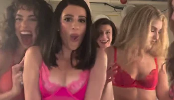 Five women are posing closely together in a playful, energetic mood. They are wearing colorful lingerie and smiling or making expressive faces, with a blurry indoor background visible behind them.