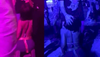 A woman in a bikini kneels on the ground in front of a man's crotch among standing people under colorful purple and blue lighting at a crowded festival.
