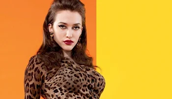 A woman with long brown hair and bold makeup is wearing a leopard print top, standing against a background split between orange and yellow.