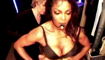 Janet Jackson wearing a headset microphone adjusts her black bra backstage, while another person stands behind her, helping with preparations. The setting appears busy and focused.