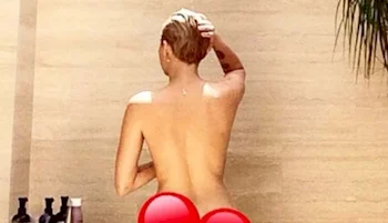 A girl with short hair stands outdoors with their back to the camera, appearing to be nude. Their lower body is covered with heart-shaped graphics. Bottles are visible on a surface nearby.