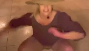 Britney Spears wearing a wide-brimmed hat and a loose outfit appears to be in mid-motion, squatting or dancing indoors on a tiled floor. The image is slightly blurred, conveying movement.