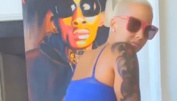 A woman with bleached blond hair, wearing sunglasses and a blue top, poses in front of a colorful painting of a person with sunglasses.