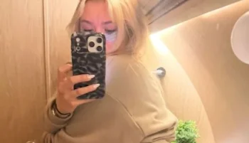 A girl with long blonde hair takes a mirror selfie in a warmly lit room, holding a phone with a patterned case. The background includes a wooden wall and a small potted plant.