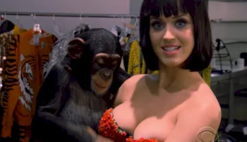 A woman with short dark hair and a red embellished top smiles at the camera while holding a chimpanzee in a backstage area with costumes hanging in the background.