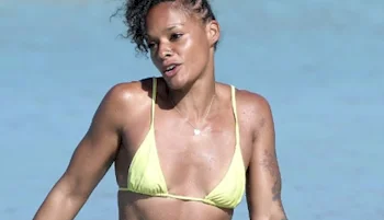 A woman wearing a yellow bikini is standing in shallow, clear blue water, with wet skin and curly hair, outdoors on a sunny day.