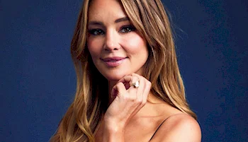 A woman with long, light brown hair poses against a dark blue background, smiling softly with her hand touching her chin. She wears a sleeveless top and a ring on her finger.