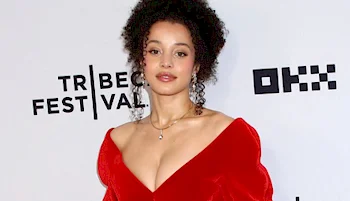 A woman with curly hair wearing a red, off-the-shoulder dress poses in front of a Tribeca Festival step-and-repeat backdrop.