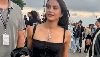 A woman with shoulder-length dark hair, wearing a black sleeveless top and silver pendant necklace, walks outdoors among a crowd of people under a cloudy sky.