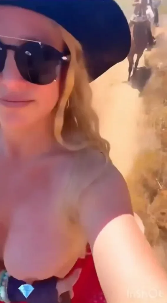 A woman wearing sunglasses and a wide-brimmed hat takes a selfie while riding a horse on a dusty trail, with other riders and horses visible in the background.