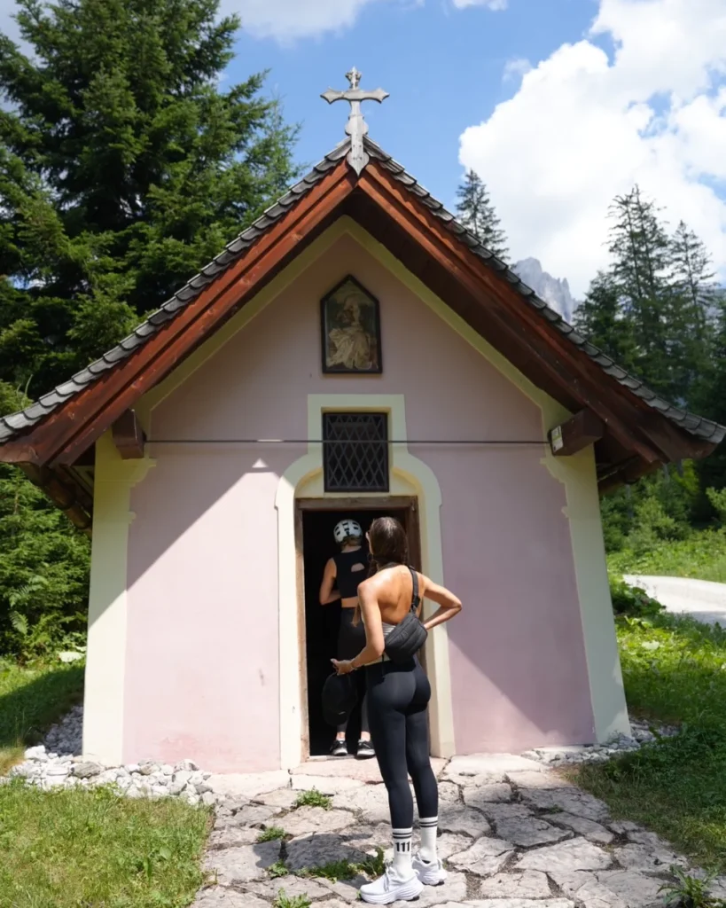 Two women in athletic clothing stand outside a small, pink chapel surrounded by trees. One woman faces the chapel, while the other stands in the doorway. The sky above is mostly clear with some clouds.
