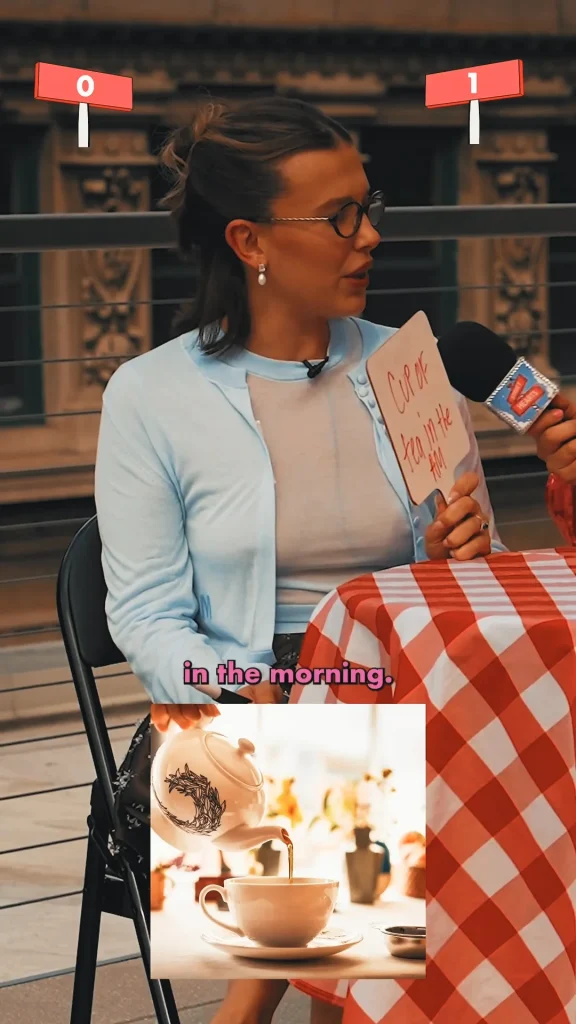 A woman sits on a chair holding a card that says Cup of tea in the AM while being interviewed. She wears glasses, earrings, and a light blue cardigan. The text in the morning, overlays an image of tea being poured.