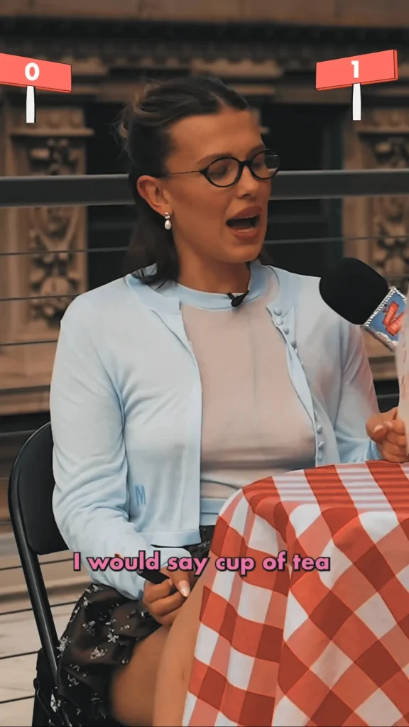 A woman with glasses wearing a light blue cardigan sits at a table with a red-and-white checkered tablecloth, speaking into a microphone. Pink text at the bottom reads, “I would say cup of tea.”.