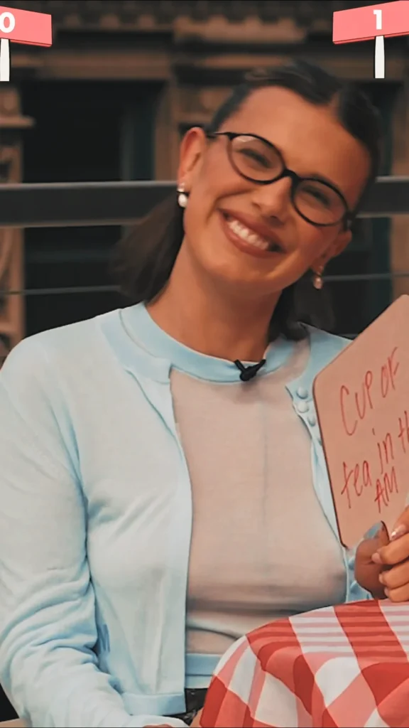 A smiling person wearing glasses and a light blue cardigan holds a whiteboard with the handwritten phrase Cup of tea in the AM. The background appears to be outdoors with buildings visible.