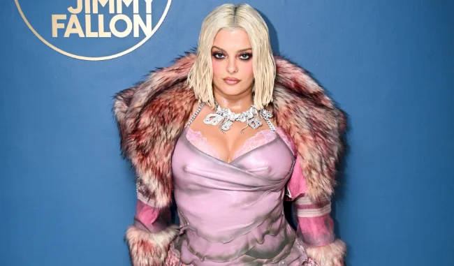 A woman with platinum blonde hair wears a pink dress, a bold pink fur coat, and a statement necklace, standing in front of a blue background with “Jimmy Fallon” written in a circle at the top left.