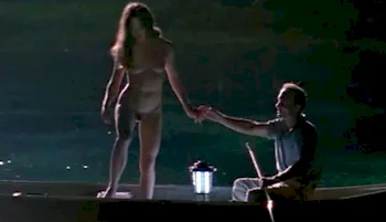 A naked woman standing on the edge of a rowboat. A man is sitting next to her and they are holding hands. She looks like she is about to jump in the water. The scene is at night.