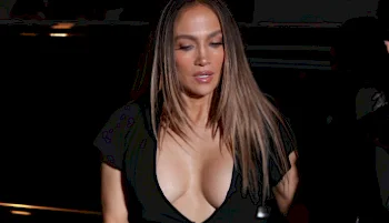 A woman with long straight hair wearing a black deep V-neck dress looks down while standing in a dimly lit setting.