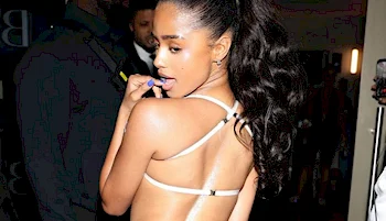 A woman with long dark hair in a ponytail, wearing a strappy open-back outfit, looks over her shoulder while brushing her teeth at an indoor event.