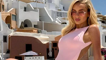 Woman in a pink swimsuit and sunglasses stands outdoors with white stucco buildings in the background, under bright sunlight.
