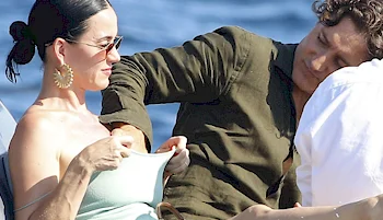 A couple sitting closely together outdoors. The woman wearing sunglasses adjusts their light-colored dress while the other, wearing a dark shirt, leans in with touching her chest. Blue water is visible in the background.