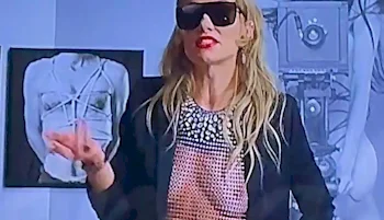 A woman with long blonde hair, wearing large black sunglasses a black jacket and sheer top, gestures with her hand. Behind her are black-and-white photos of a restrained person and vintage camera equipment.
